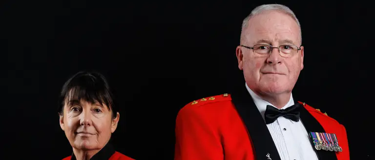 Two military personnel in formal red dress uniforms with medals stand against a black background.