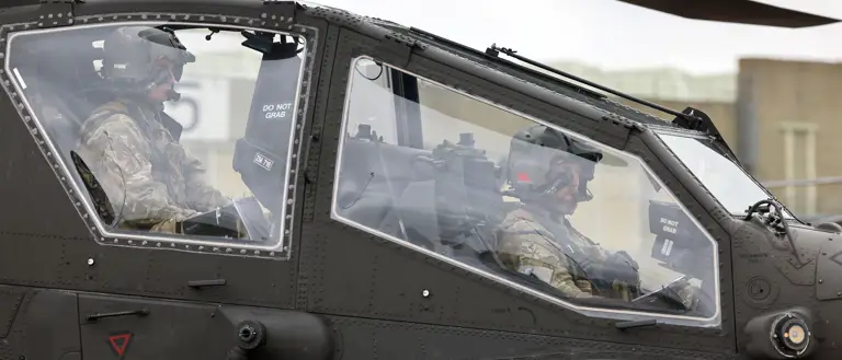 Two soldiers in full combat gear seated inside a military attack helicopter cockpit preparing for flight.