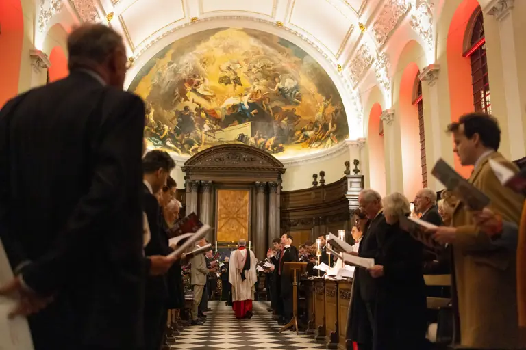 Congregation standing and holding hymnals inside a grand church with a large religious mural above the altar.