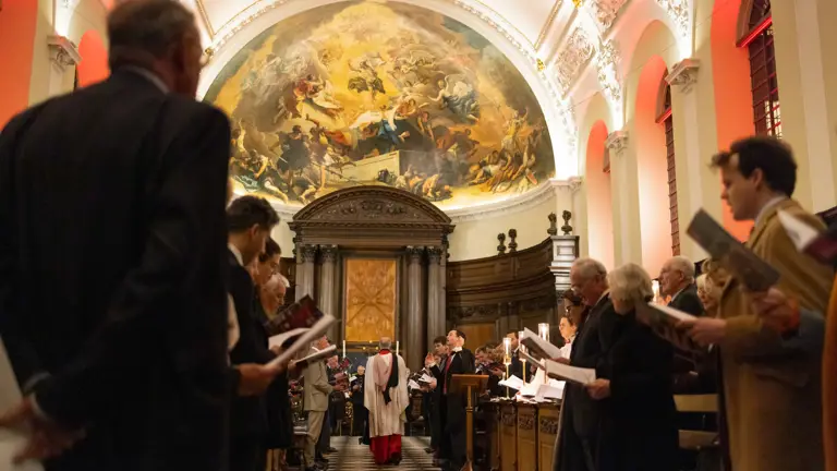 Congregation standing and holding hymnals inside a grand church with a large religious mural above the altar.