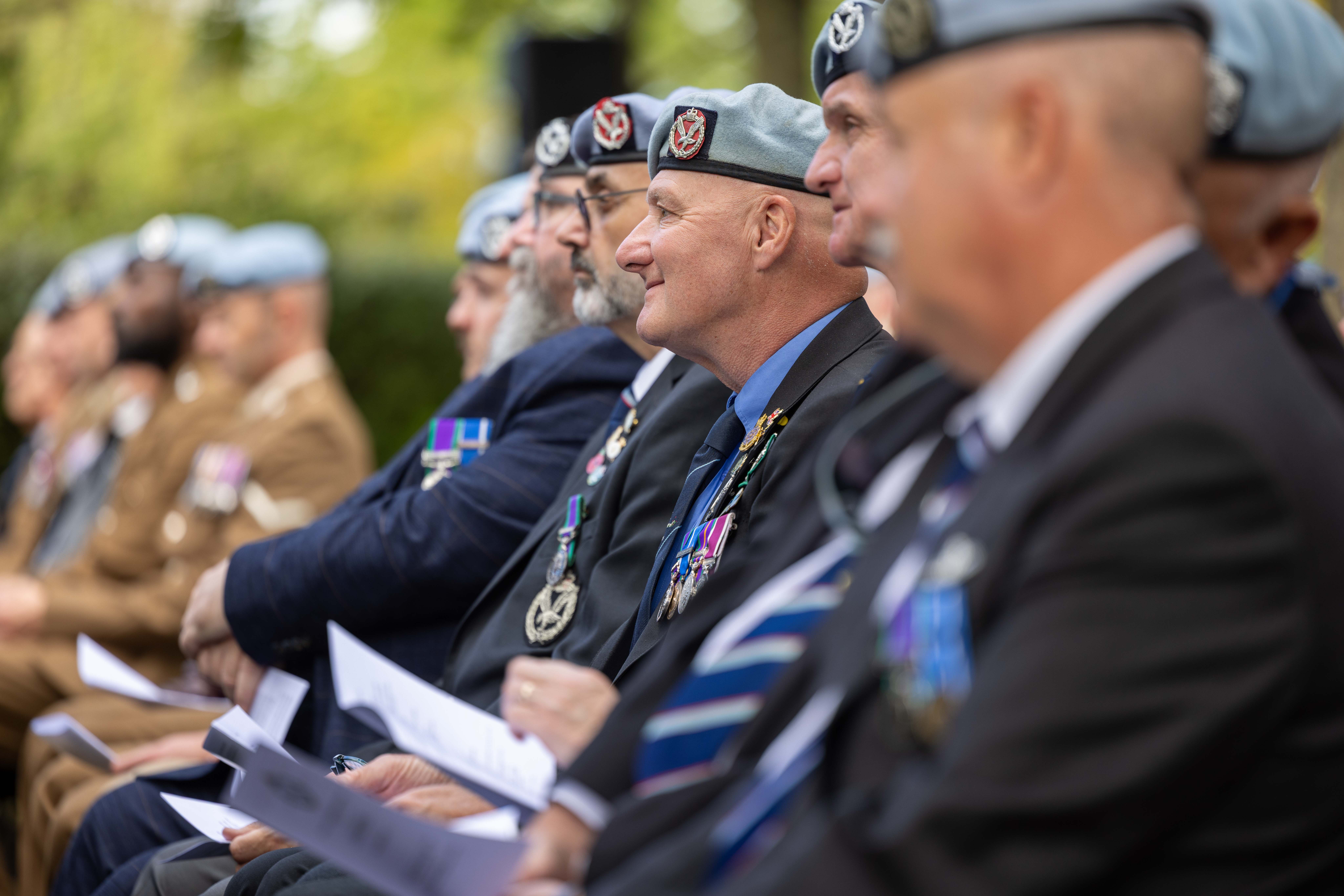 A group of people wearing uniforms and blue berets are seated in an outdoor setting. They have medals on their jackets and are holding papers, appearing to participate in a formal event. The background features greenery.