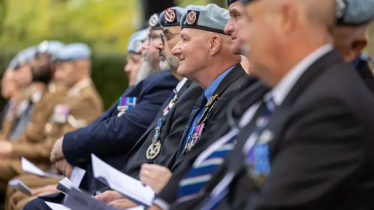 A group of people wearing uniforms and blue berets are seated in an outdoor setting. They have medals on their jackets and are holding papers, appearing to participate in a formal event. The background features greenery.
