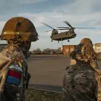 Soldiers wearing their camouflage uniform with a red and blue flash on their arm with their paratrooper wings above it stand back as a chinook coming into be loaded.