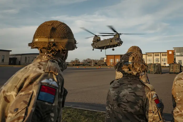 Soldiers wearing their camouflage uniform with a red and blue flash on their arm with their paratrooper wings above it stand back as a chinook coming into be loaded.