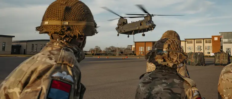 Soldiers wearing their camouflage uniform with a red and blue flash on their arm with their paratrooper wings above it stand back as a chinook coming into be loaded.