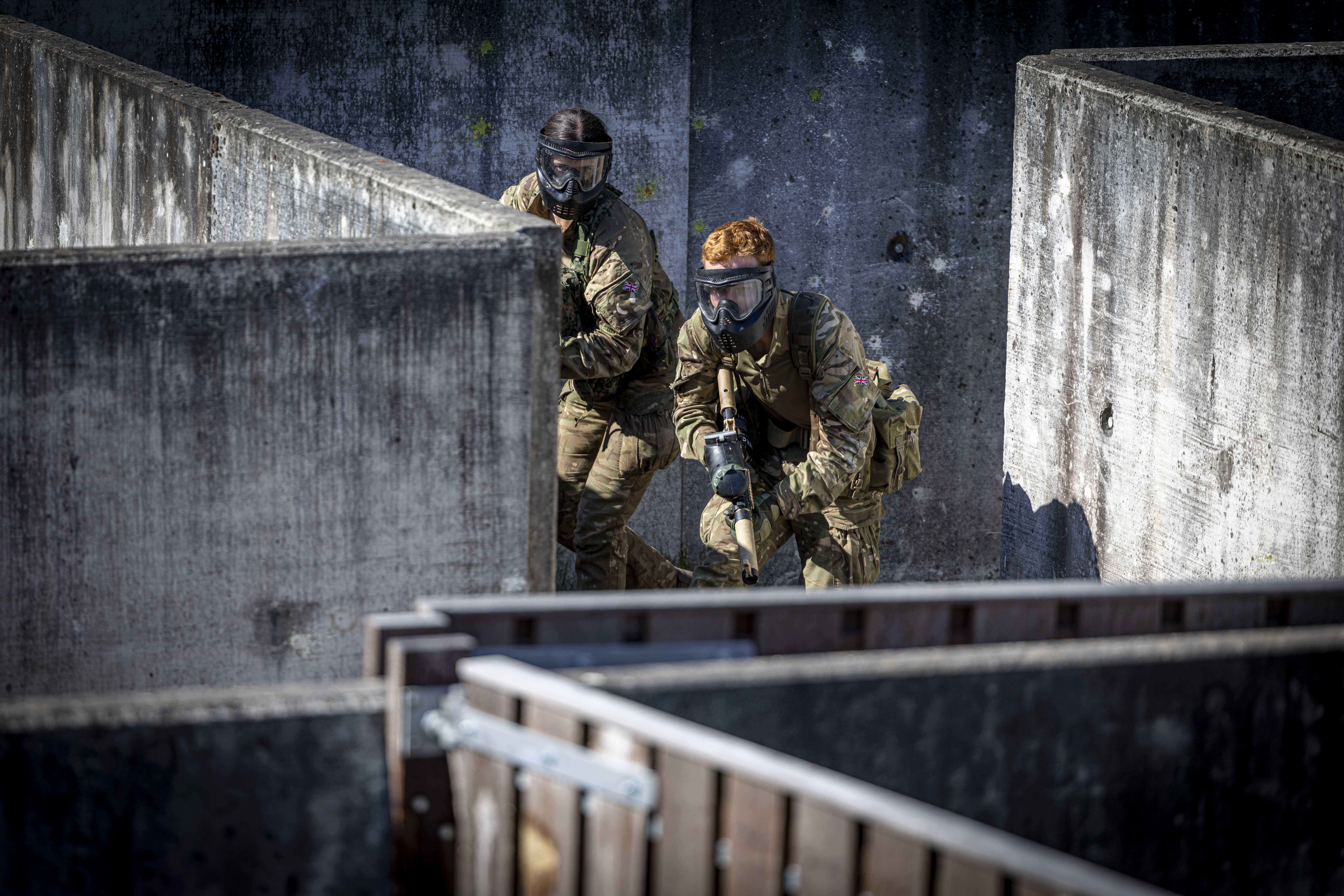 Two individuals wearing tactical gear and face protection navigate a concrete maze-like structure, possibly during a training exercise or simulation.