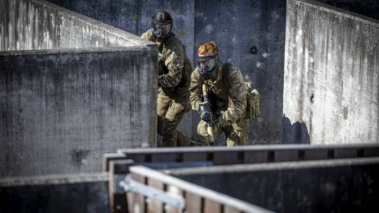 Two individuals wearing tactical gear and face protection navigate a concrete maze-like structure, possibly during a training exercise or simulation.