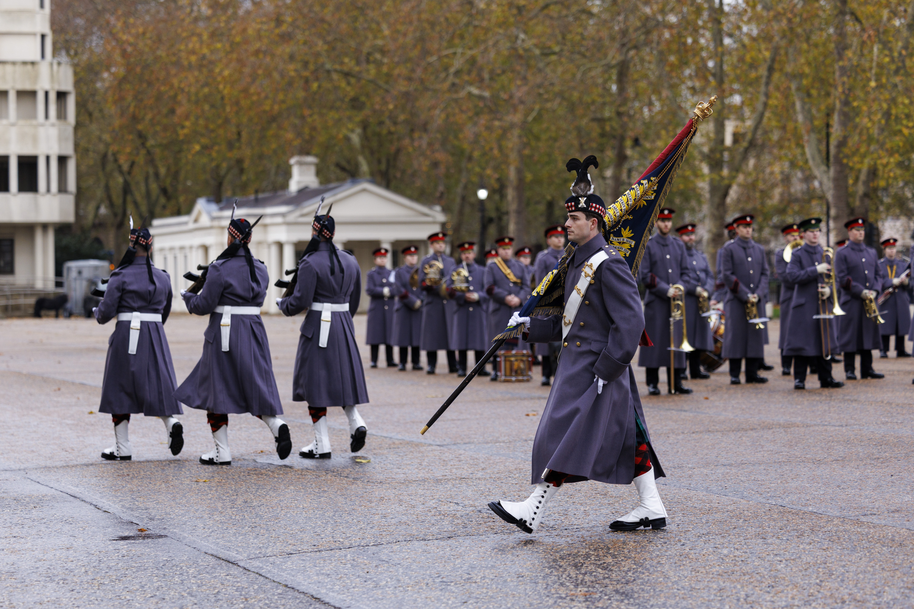 A military band in dark grey uniforms march in formation during a ceremonial event outdoors.