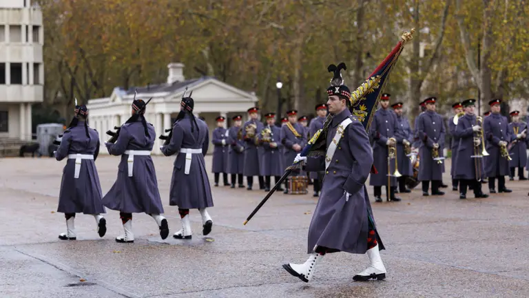 A military band in dark grey uniforms march in formation during a ceremonial event outdoors.