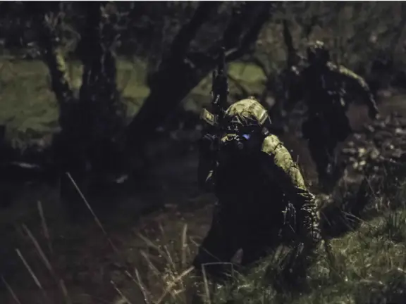 A dark image of a soldier in the jungle carrying a gun and wearing green camouflage and night vision goggles.
