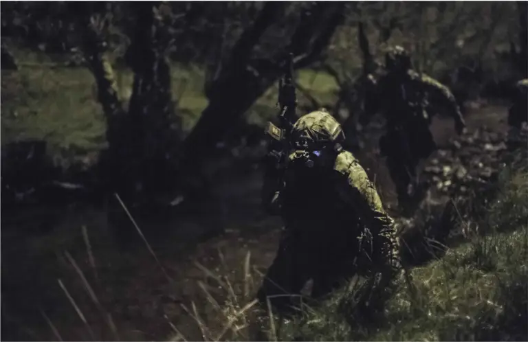 A dark image of a soldier in the jungle carrying a gun and wearing green camouflage and night vision goggles.