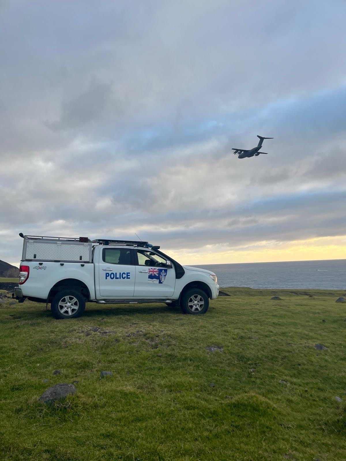 A police truck is parked on a grassy hill overlooking the ocean under a cloudy sky. A large aircraft flies overhead, creating a dramatic scene.
