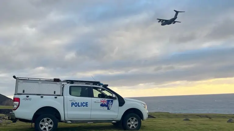 A police truck is parked on a grassy hill overlooking the ocean under a cloudy sky. A large aircraft flies overhead, creating a dramatic scene.
