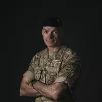 A man stands with his arms folded across his chest for a portrait. He is wearing camouflage uniform and a dark blue beret.