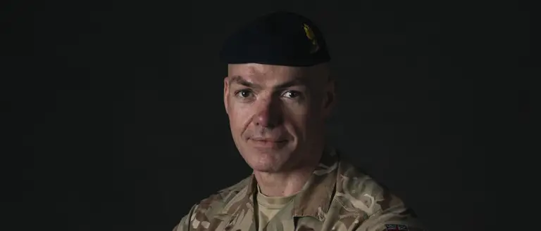 A man stands with his arms folded across his chest for a portrait. He is wearing camouflage uniform and a dark blue beret.