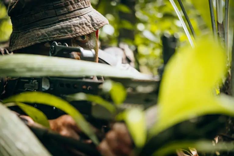 Soldier camouflaged in dense jungle foliage aiming a rifle with focused eyes visible under a boonie hat.