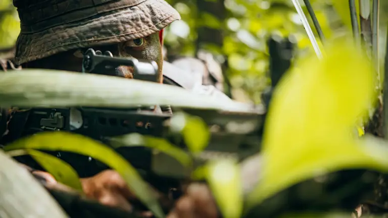 Soldier camouflaged in dense jungle foliage aiming a rifle with focused eyes visible under a boonie hat.