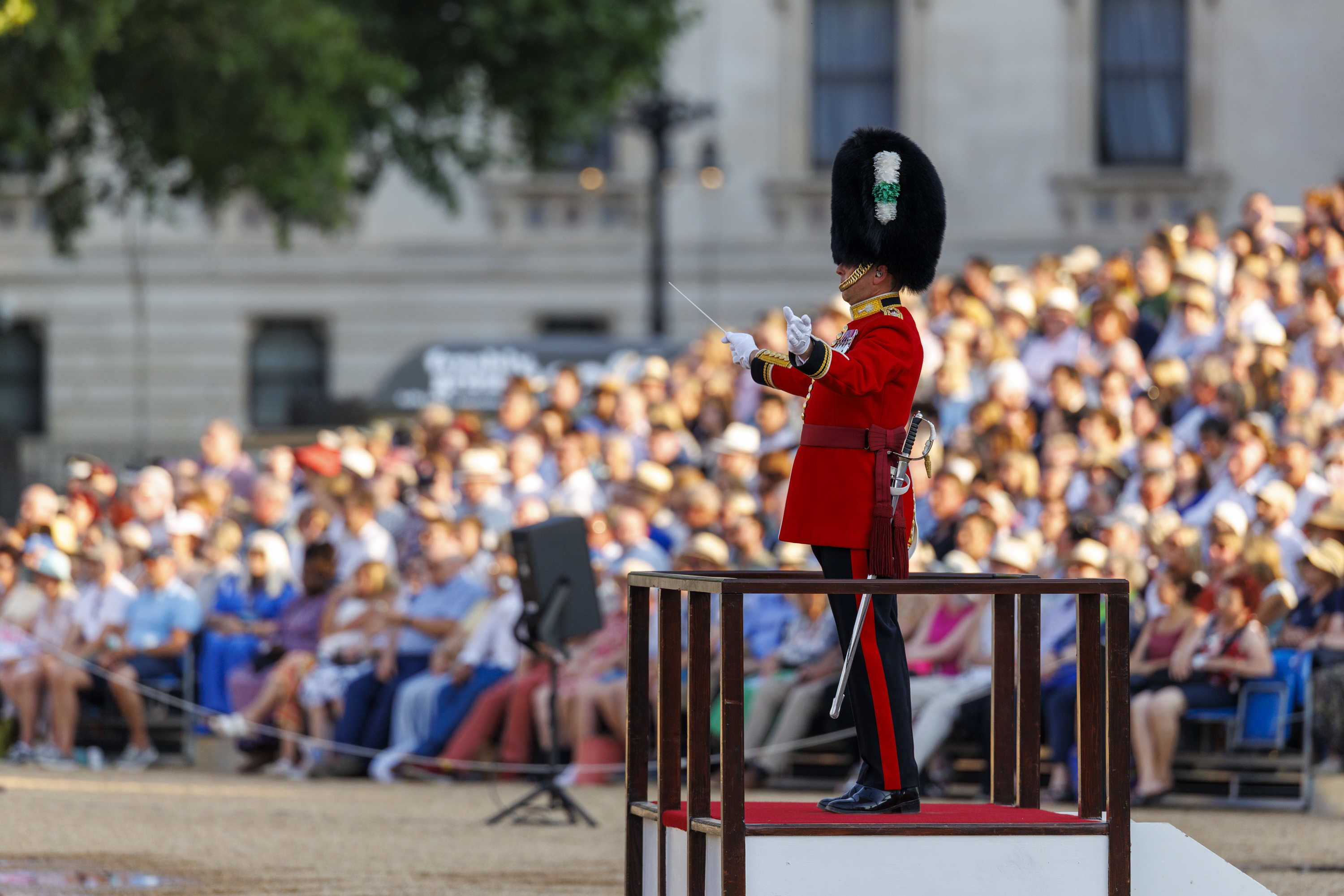 A person in a red uniform and tall bearskin hat stands on a platform, conducting a performance. The background shows a large, seated audience outside, likely at a parade or ceremonial event.