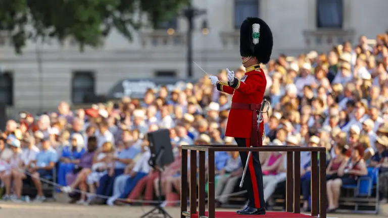 A person in a red uniform and tall bearskin hat stands on a platform, conducting a performance. The background shows a large, seated audience outside, likely at a parade or ceremonial event.
