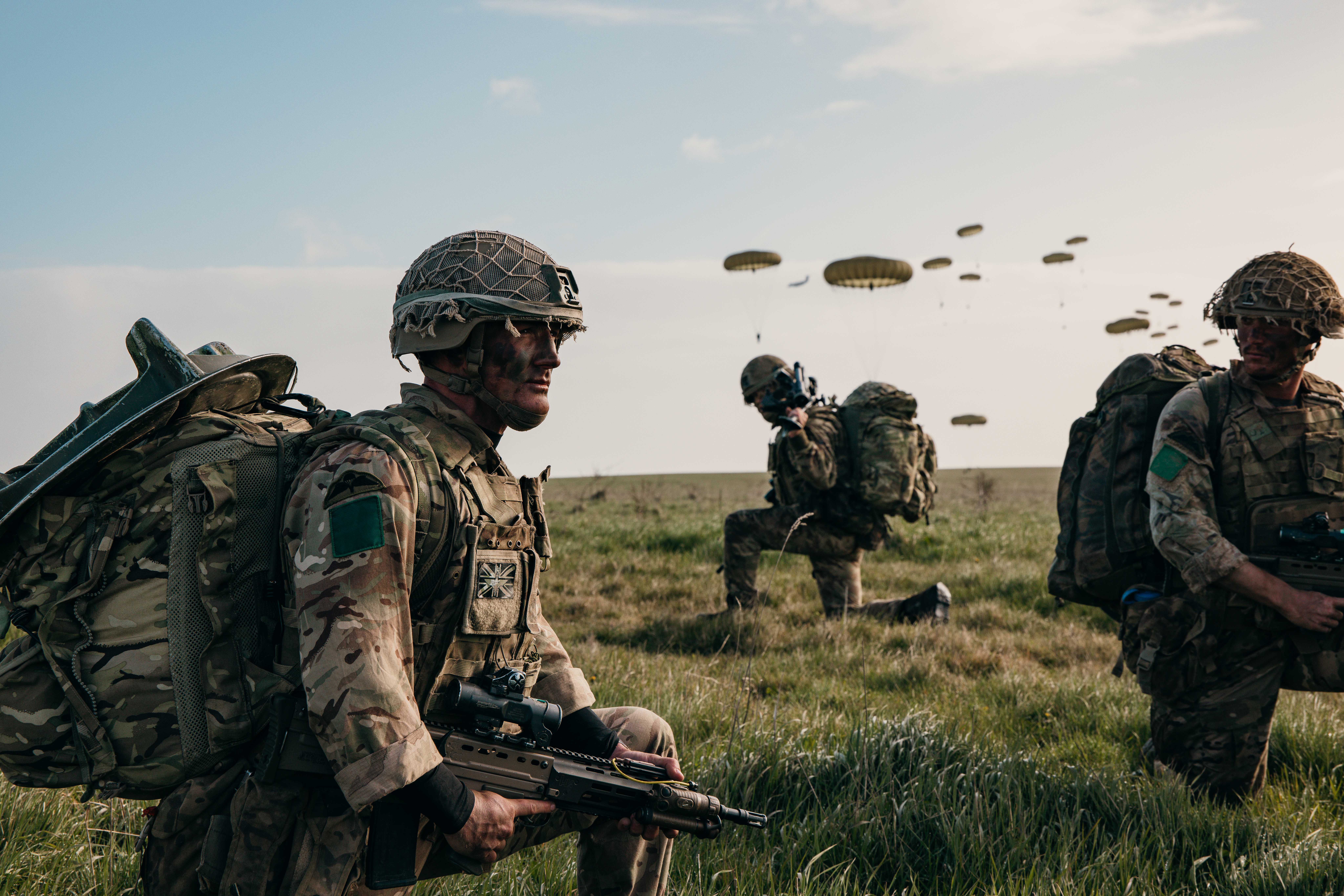 A group of soldiers in camouflage gear and helmets are on a grassy field. One is crouching with a weapon. 