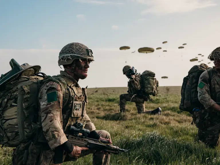 A group of soldiers in camouflage gear and helmets are on a grassy field. One is crouching with a weapon.