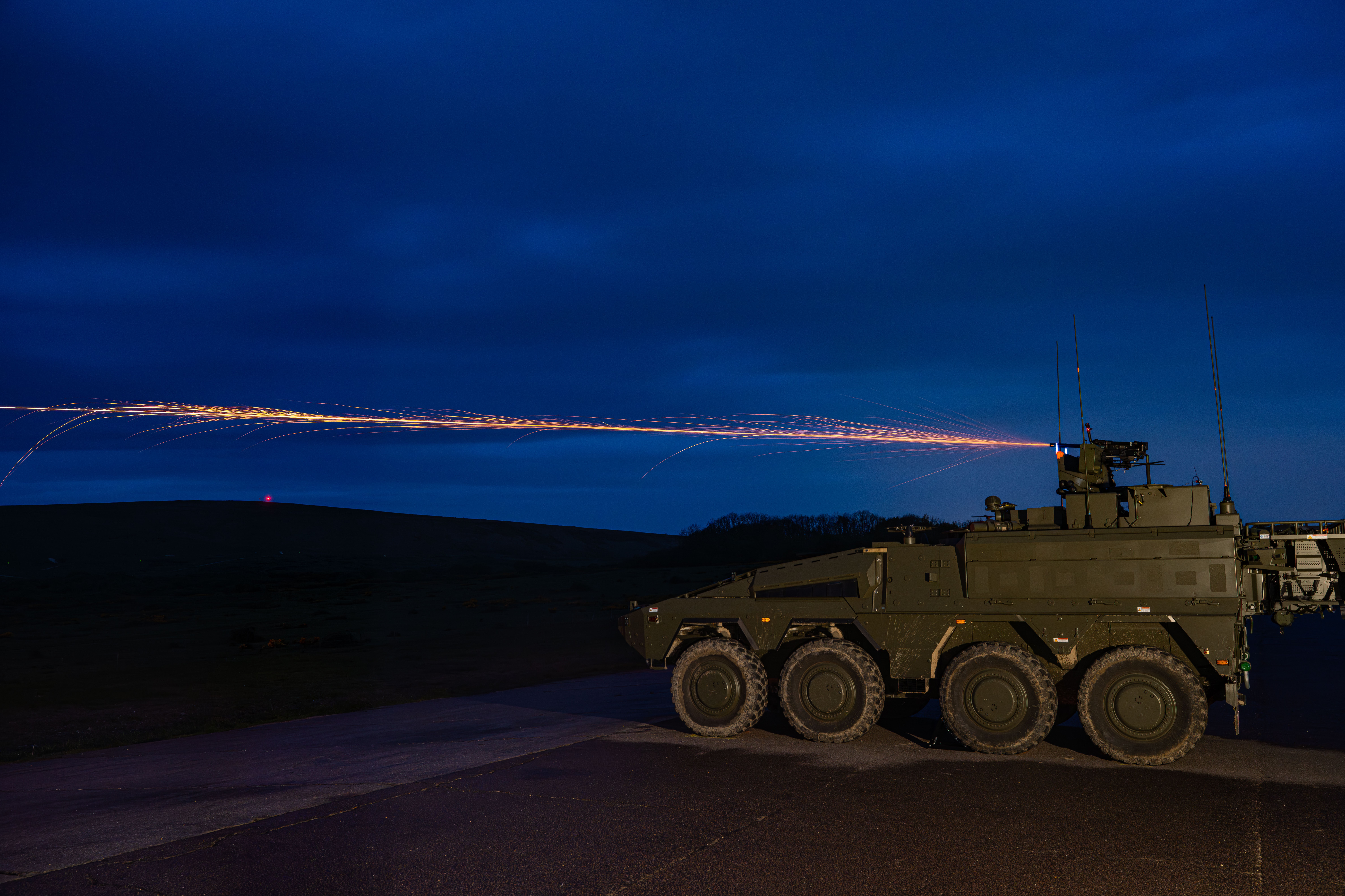 A military armoured vehicle on a road fires a tracer round into the night sky.