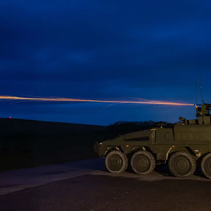 A military armoured vehicle on a road fires a tracer round into the night sky.