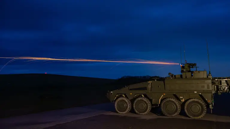 A military armoured vehicle on a road fires a tracer round into the night sky.