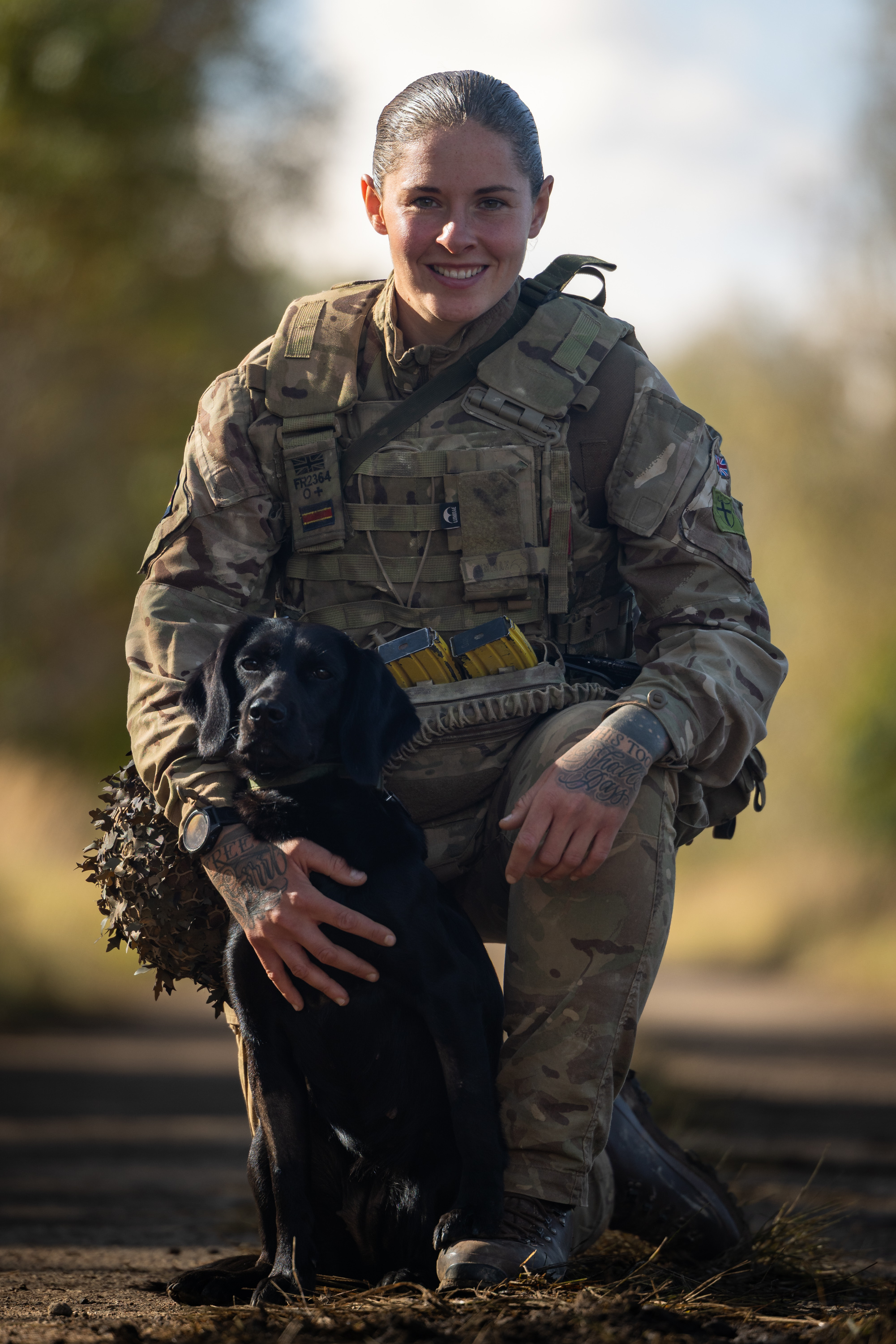 Soldier in full camouflage gear kneeling outdoors with a black dog sitting calmly beside them on a dirt path.