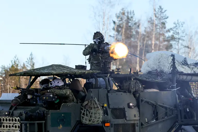 Soldier firing a mounted machine gun from an armored military vehicle in a forested area during daylight.