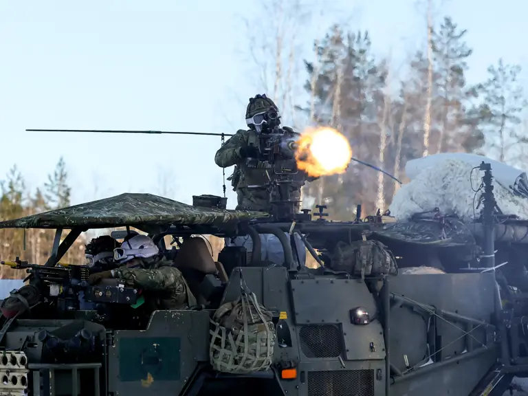 Soldier firing a mounted machine gun from an armored military vehicle in a forested area during daylight.