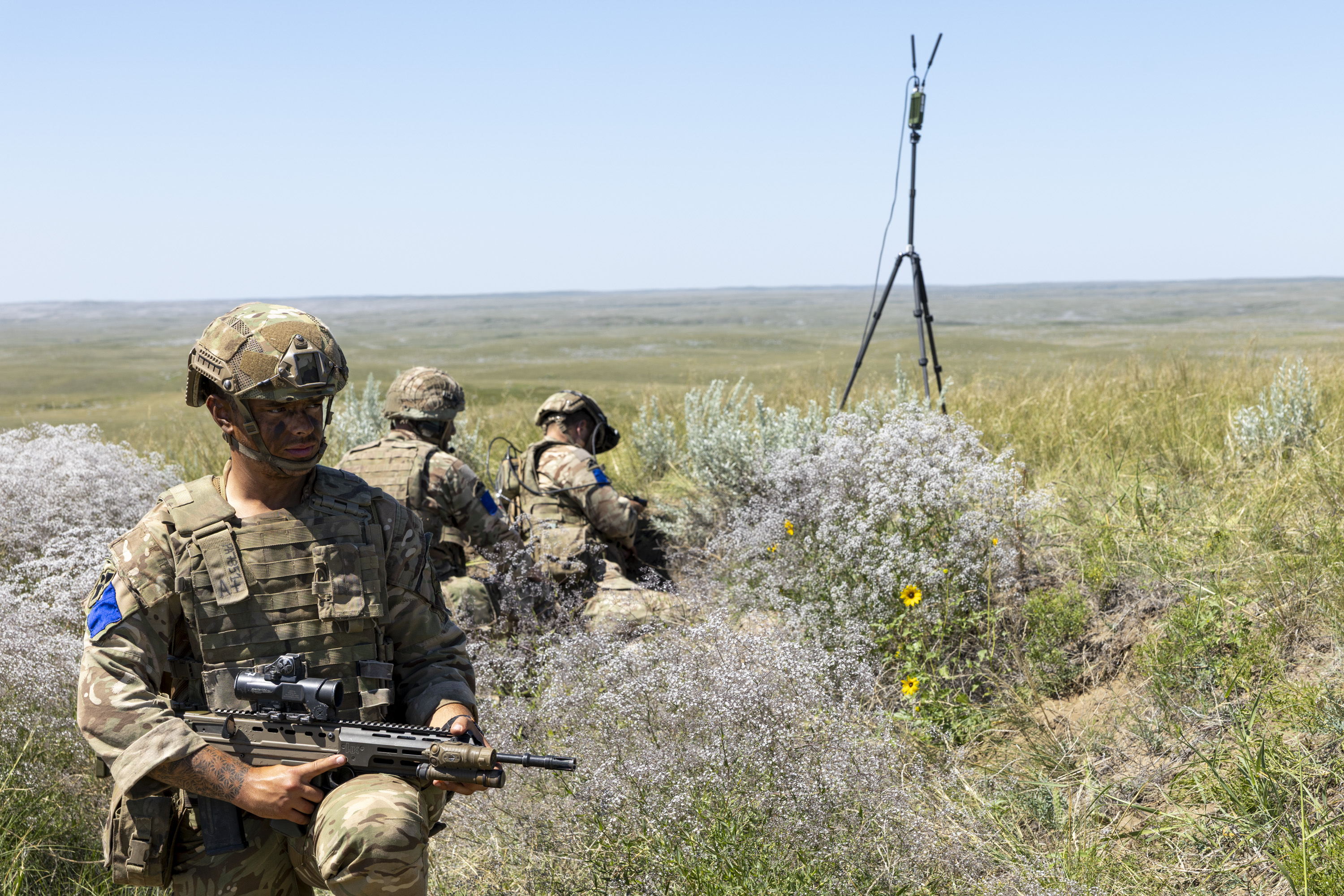 Soldiers in camouflage uniforms are positioned in tall grassy terrain during a military exercise. One soldier is in the foreground holding a rifle, with others crouched in the background near communication equipment. The landscape is spacious and open with a clear blue sky.