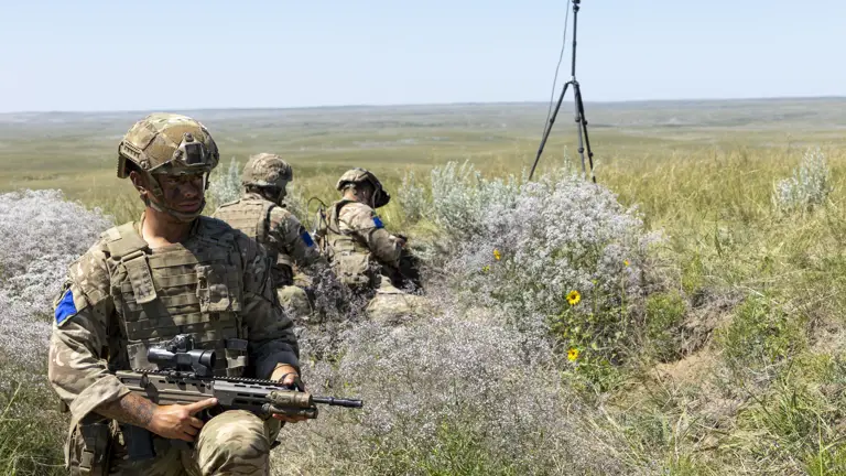 Soldiers in camouflage uniforms are positioned in tall grassy terrain during a military exercise. One soldier is in the foreground holding a rifle, with others crouched in the background near communication equipment. The landscape is spacious and open with a clear blue sky.