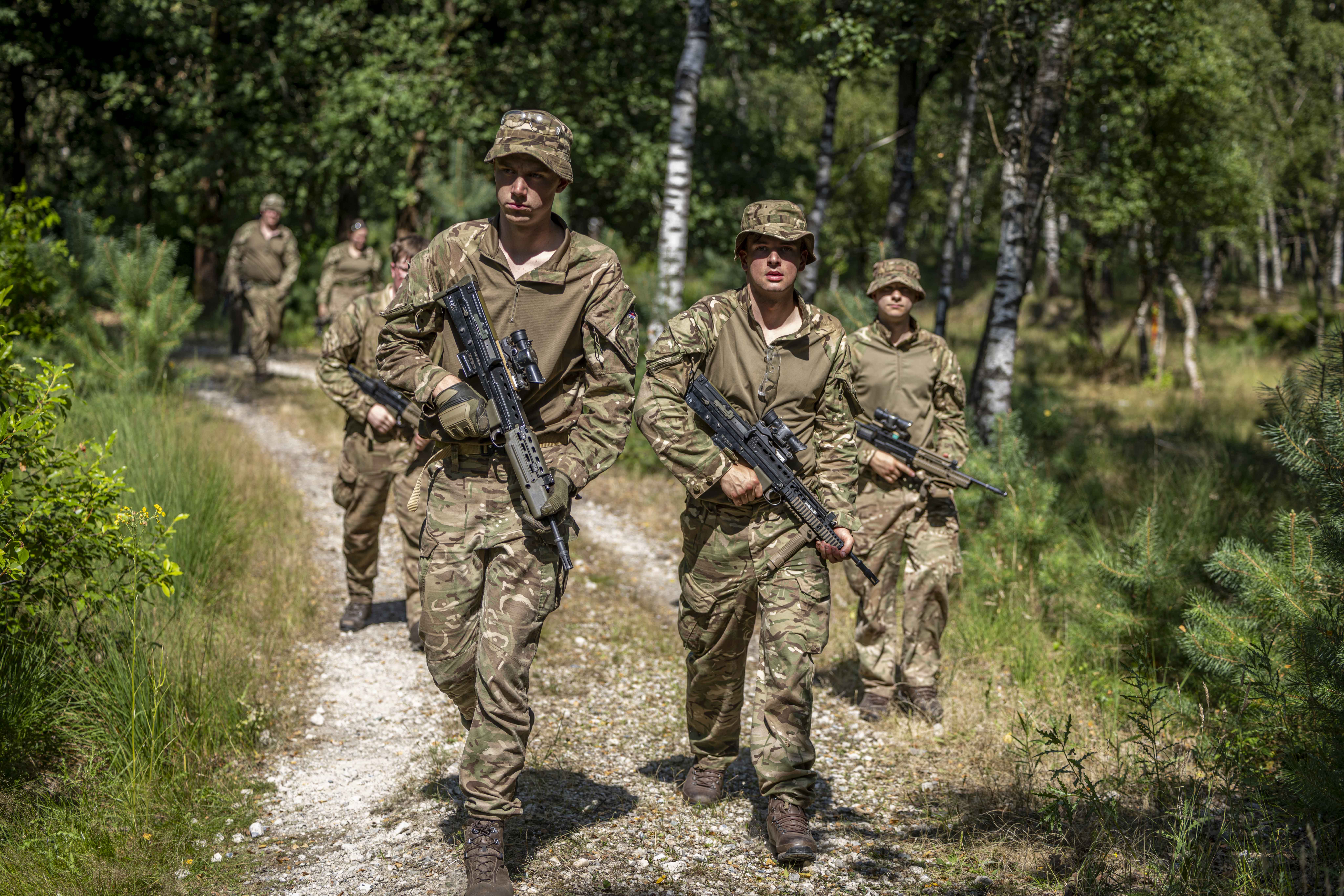 Military personnel in camouflage uniforms walk along a forest path, carrying rifles. They are in a wooded area with green trees and grass on a sunny day.