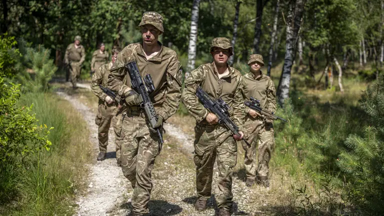 Military personnel in camouflage uniforms walk along a forest path, carrying rifles. They are in a wooded area with green trees and grass on a sunny day.