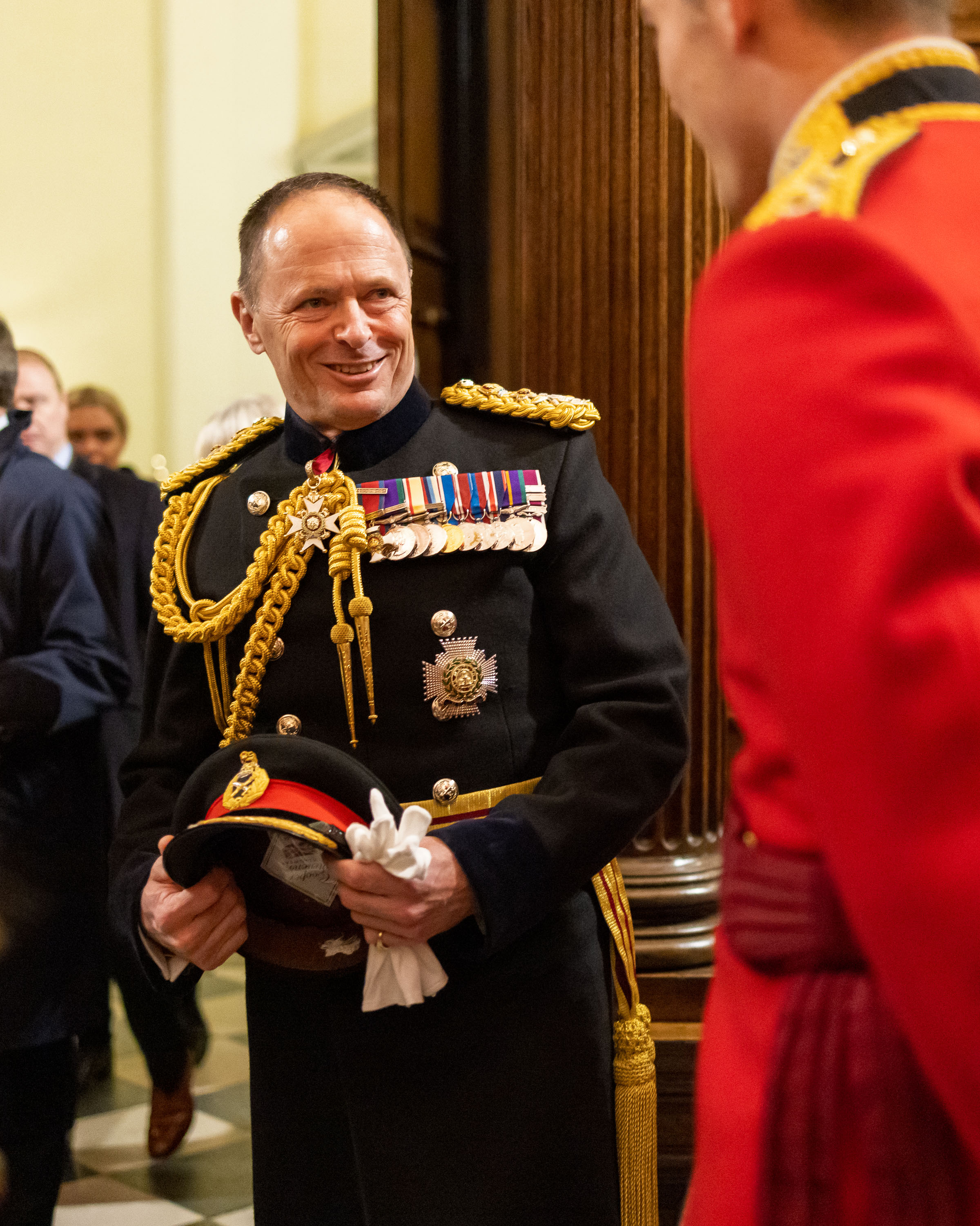 Military officer in formal dress uniform holding a ceremonial hat during an indoor event.