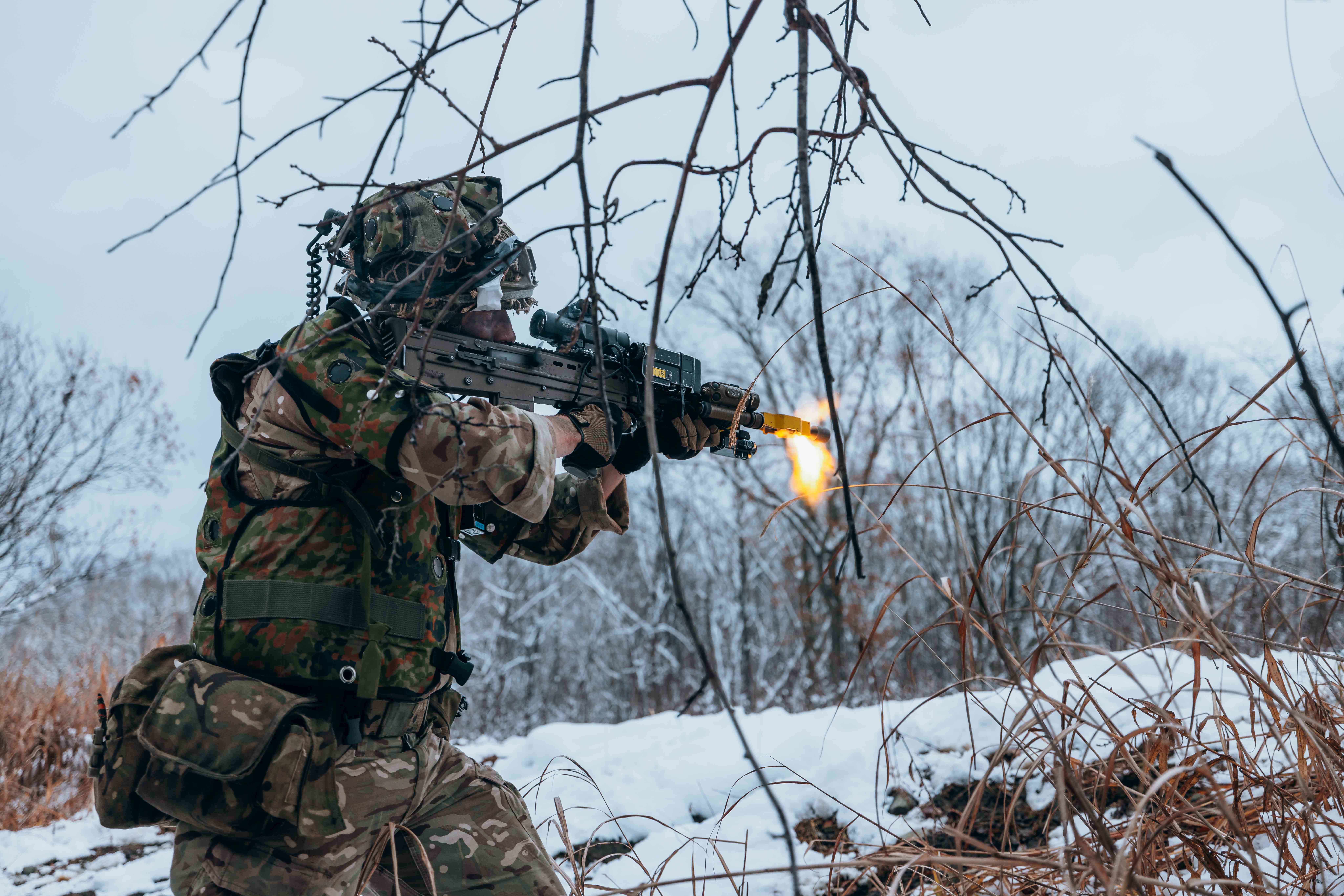 Soldier in camouflage firing a rifle in a snowy forest with bare trees around.