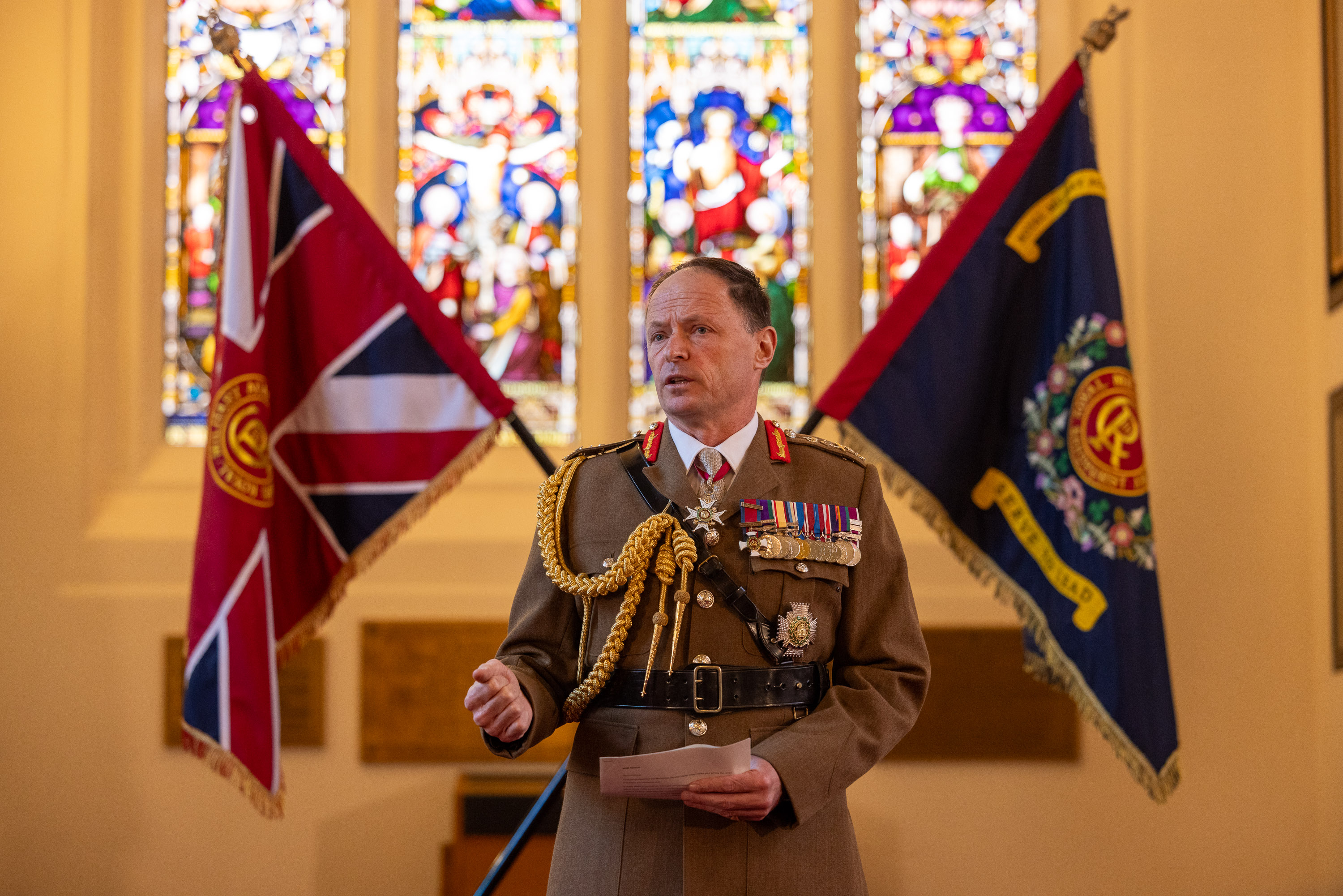 man in brown unifrom decorated with medals stands in a church style room giving a speak with two flags crossed behind him