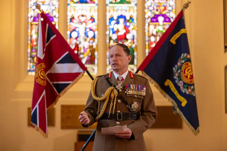 man in brown unifrom decorated with medals stands in a church style room giving a speak with two flags crossed behind him