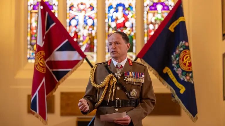 man in brown unifrom decorated with medals stands in a church style room giving a speak with two flags crossed behind him