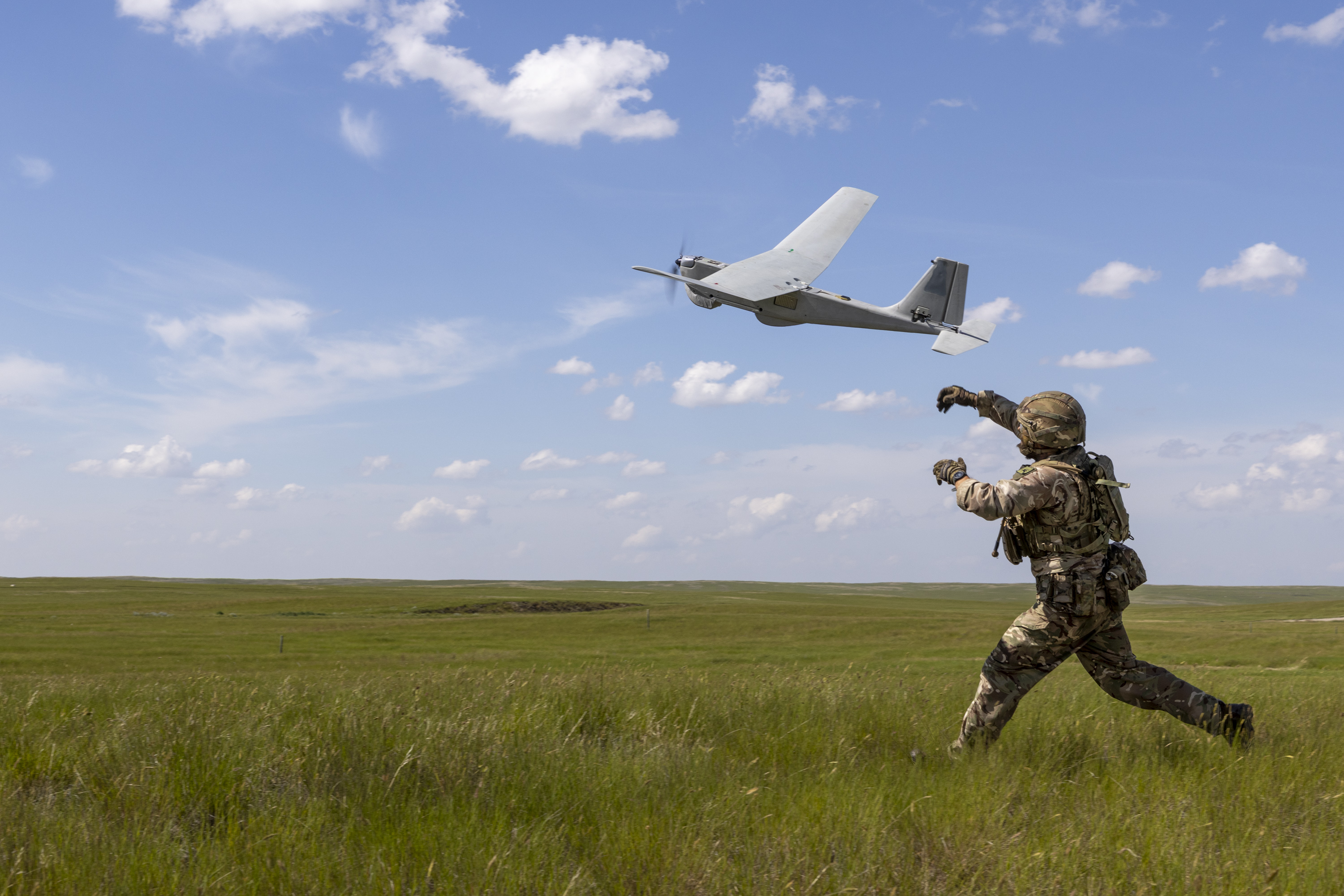 A person in military gear is launching a drone into a clear blue sky, standing on open grassy terrain.