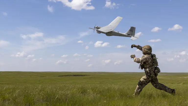 A person in military gear is launching a drone into a clear blue sky, standing on open grassy terrain.