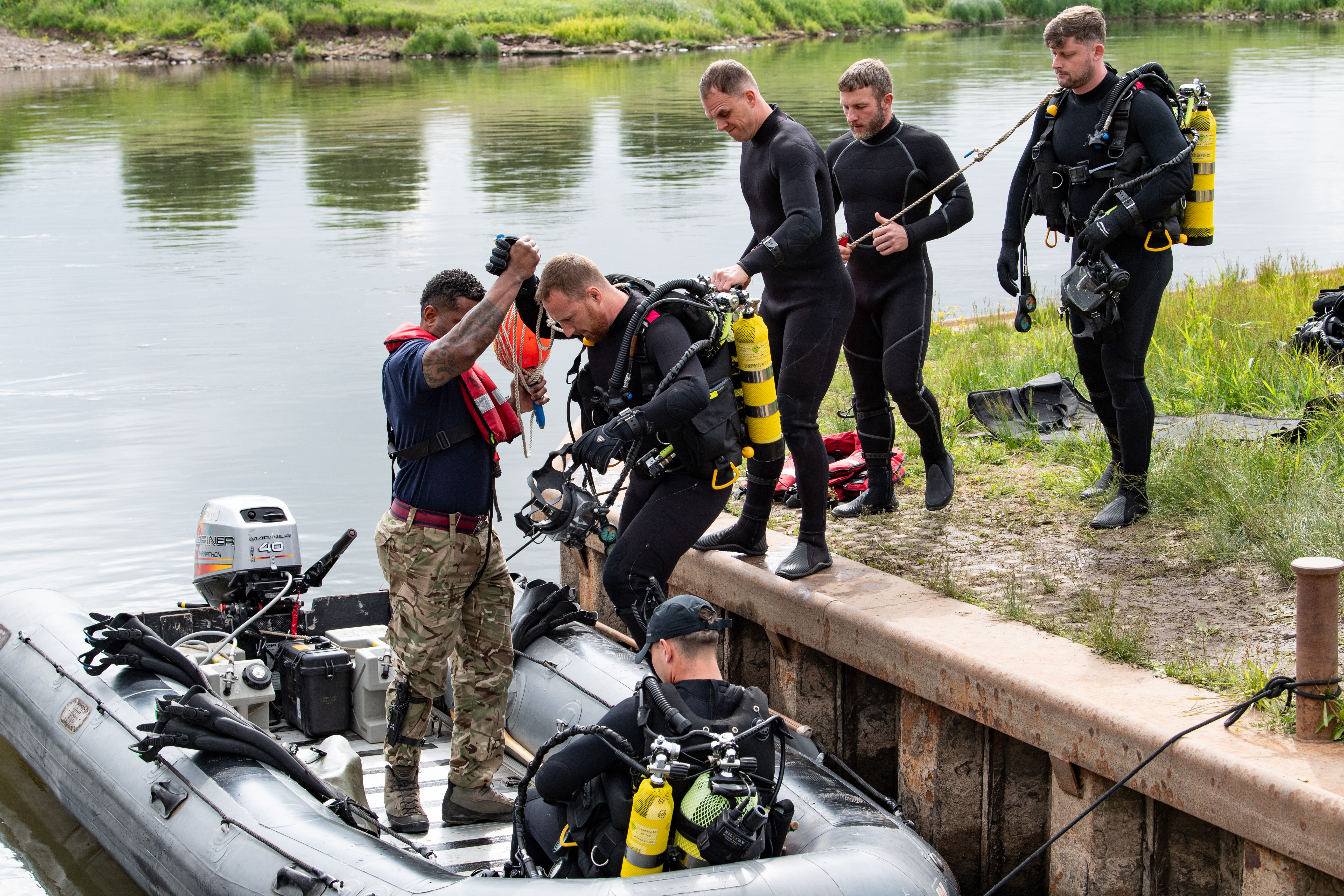 Divers in full gear preparing to board an inflatable boat by a calm riverbank with grassy surroundings.