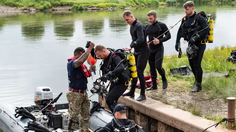 Divers in full gear preparing to board an inflatable boat by a calm riverbank with grassy surroundings.
