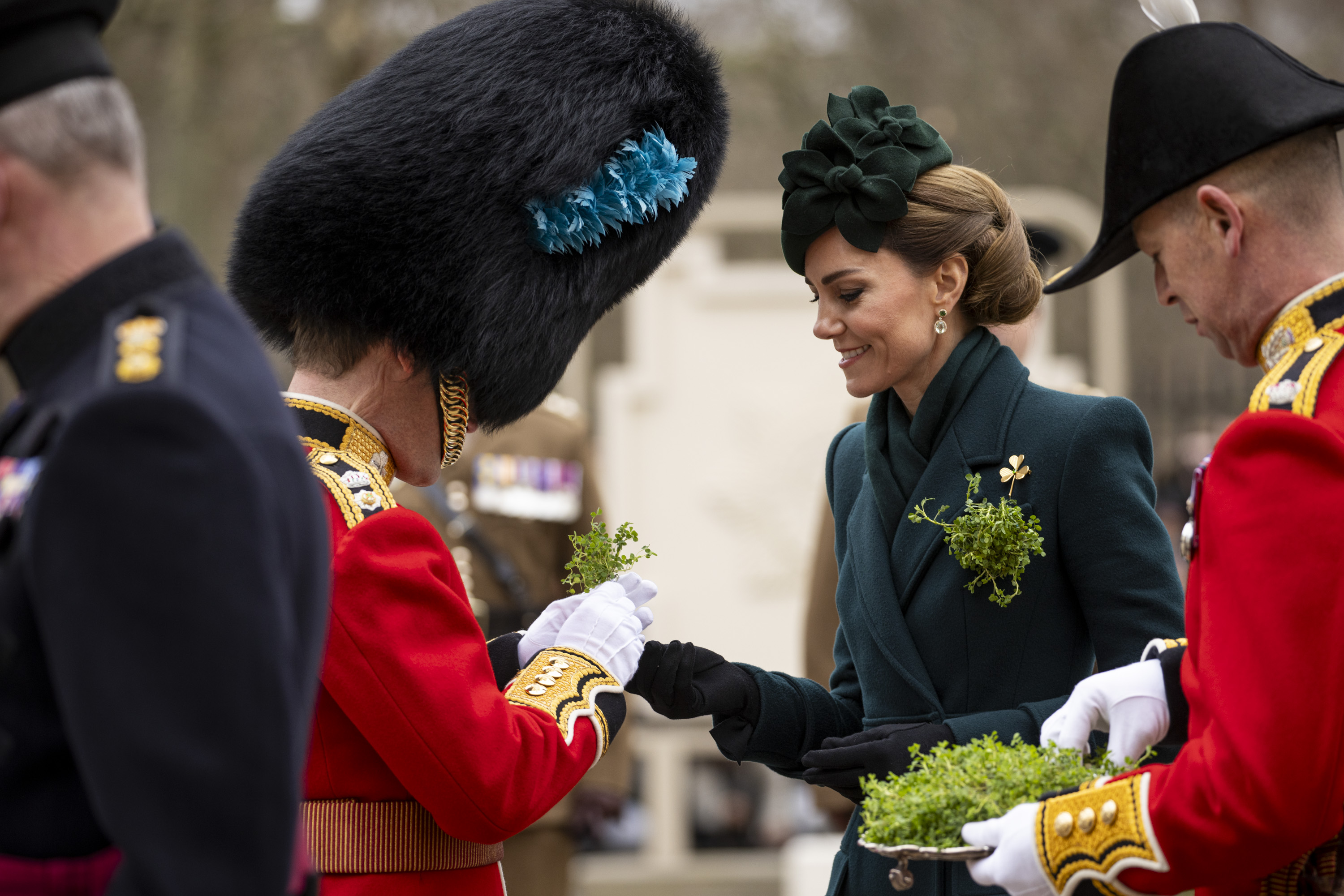 Her Royal Highness The Princess of Wales smiles as she hands a shamrock to a man in a red tunic with a bearskin hat.