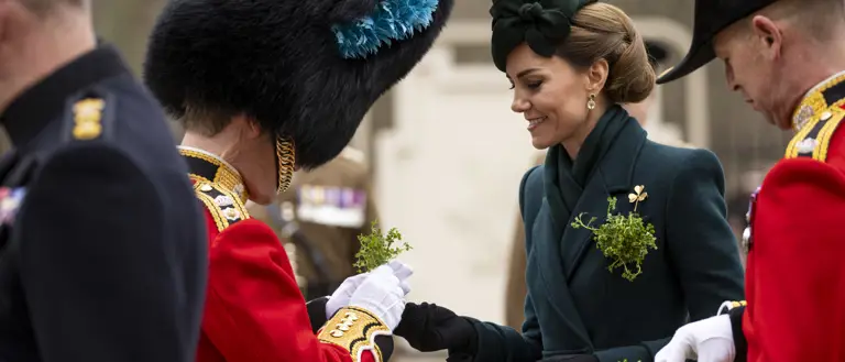 Her Royal Highness The Princess of Wales smiles as she hands a shamrock to a man in a red tunic with a bearskin hat.