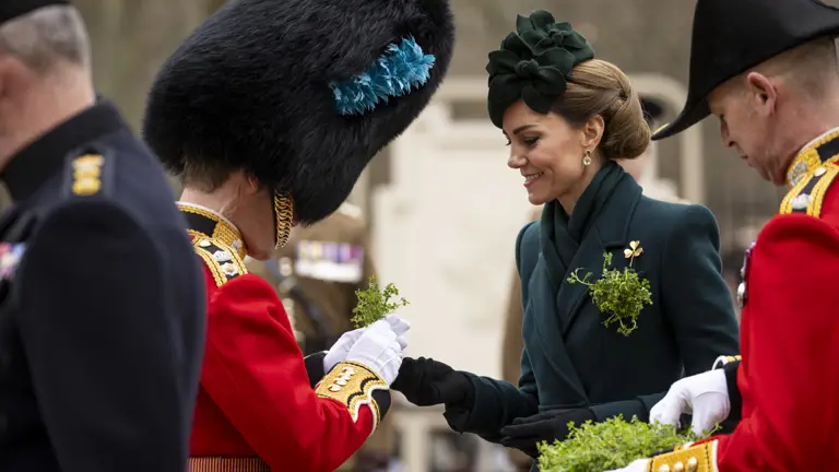 Her Royal Highness The Princess of Wales smiles as she hands a shamrock to a man in a red tunic with a bearskin hat.
