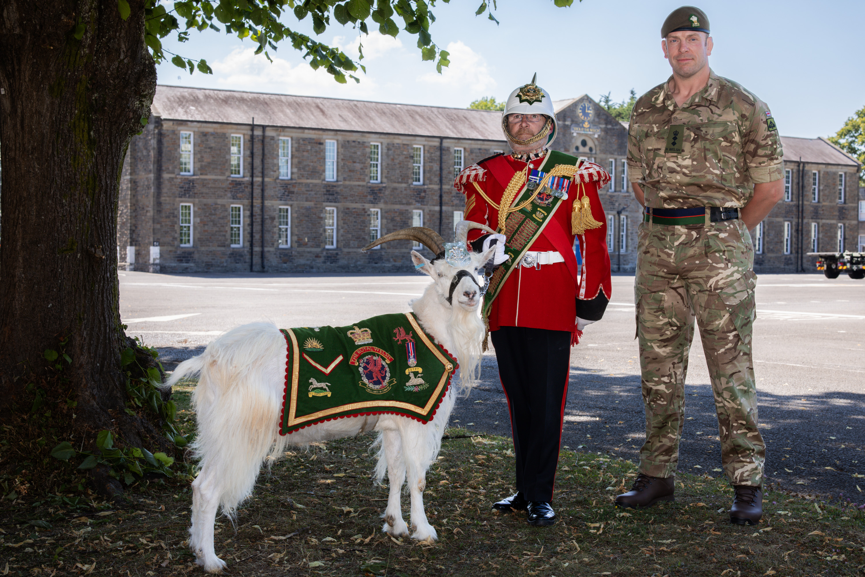 Two men stand in front of a builidng. The one on the left is holding the lead of a goat who is wearing a green ceremonial coat. The man on the left is wearing a red tunic and ceremonial helmet, the man on the right is wearing a green camouflage uniform.