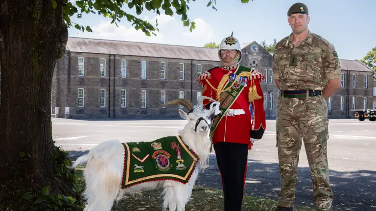 Two men stand in front of a builidng. The one on the left is holding the lead of a goat who is wearing a green ceremonial coat. The man on the left is wearing a red tunic and ceremonial helmet, the man on the right is wearing a green camouflage uniform.