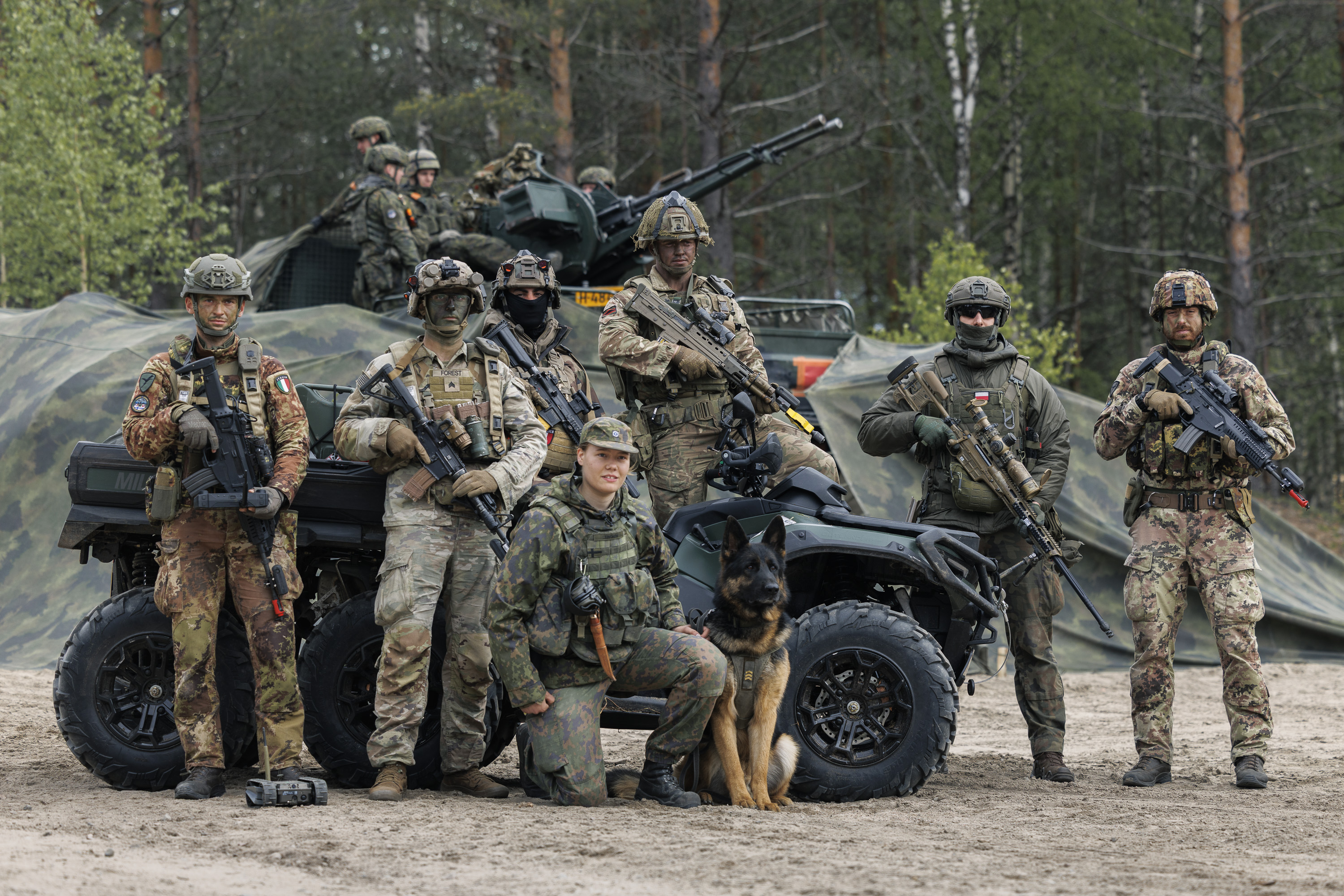 A group of military personnel in camouflage gear stands in front of a forest backdrop, posing with a K9 dog and an armored vehicle. The equipment includes rifles and a military ATV. The individuals have helmets and tactical gear, with trees and greenery in the background.